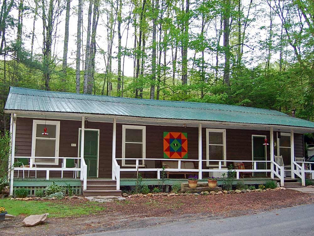 A brown cabin at Healing Springs with a green metal roof and a long front porch, featuring a colorful quilt block hanging on the wall between windows, set against a background of tall trees.