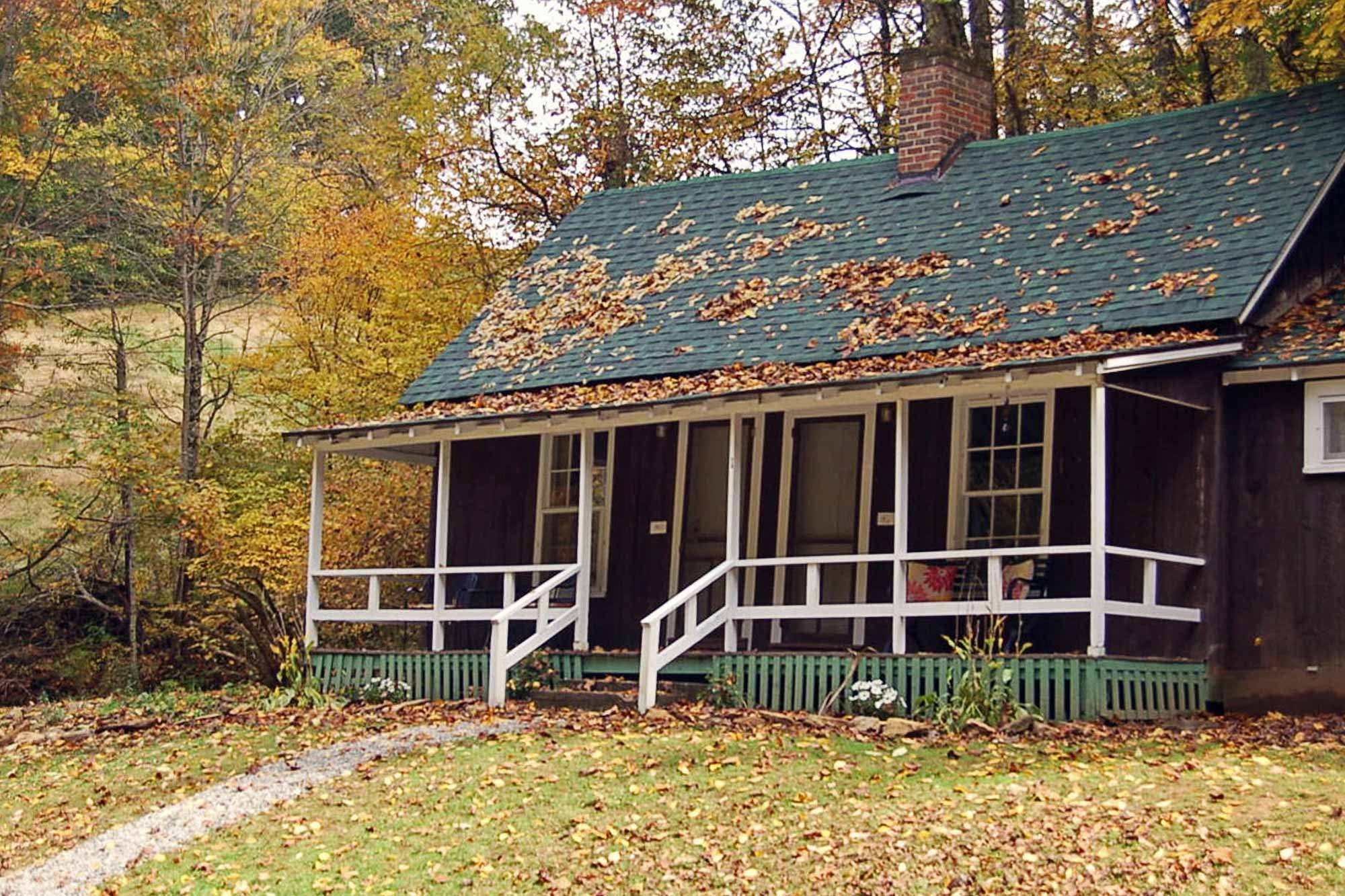 A dark wood cabin, one of the affordable cabins in Ashe County NC, with a green roof covered in fallen autumn leaves, featuring a white front porch and steps.