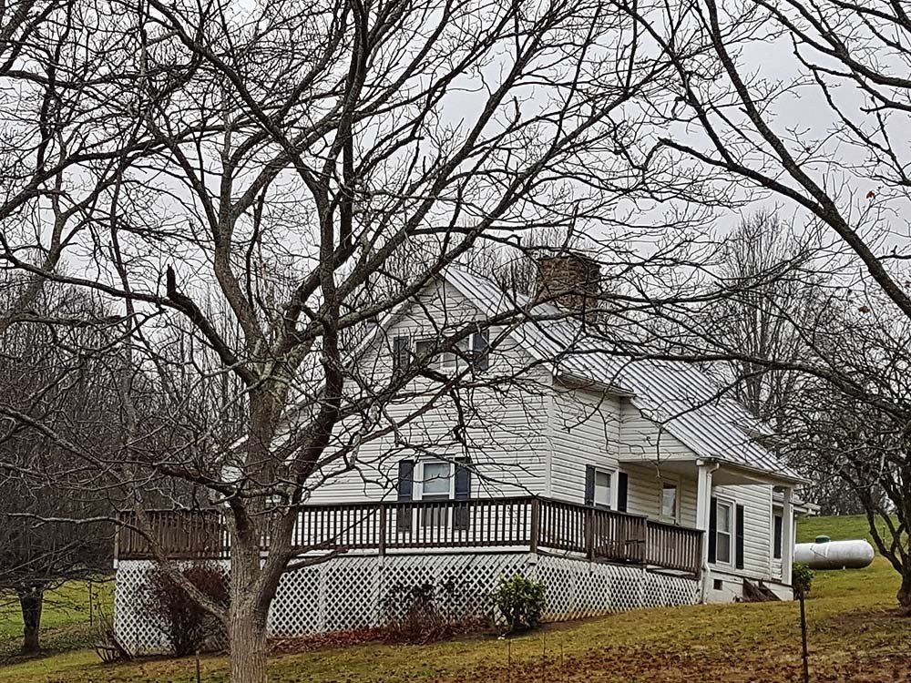 A two-story white family cabin in Ashe County, NC, featuring a metal roof and a deck, is partially obscured by bare trees in late autumn.
