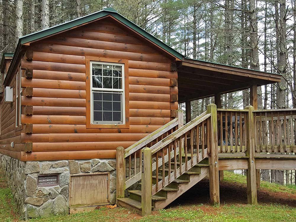 A log cabin with a stone foundation and a covered wooden deck accessed by stairs in a wooded area, typical of family cabins in Ashe County NC.