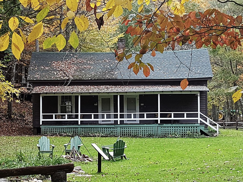 A dark, one-story cabin in Ashe County, NC, featuring a long porch with a white railing, sits on a grassy lawn with several chairs in front.